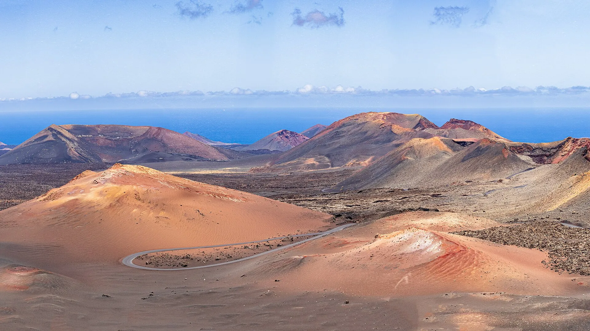 Red and ochre volcanoes in Timanfaya National Park, with a road winding between them and the sea beyond